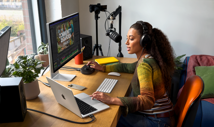Жена в дома си, която работи на бюрото си с MacBook Pro, свързан към два външни дисплея, със слушалки и която говори в микрофон.