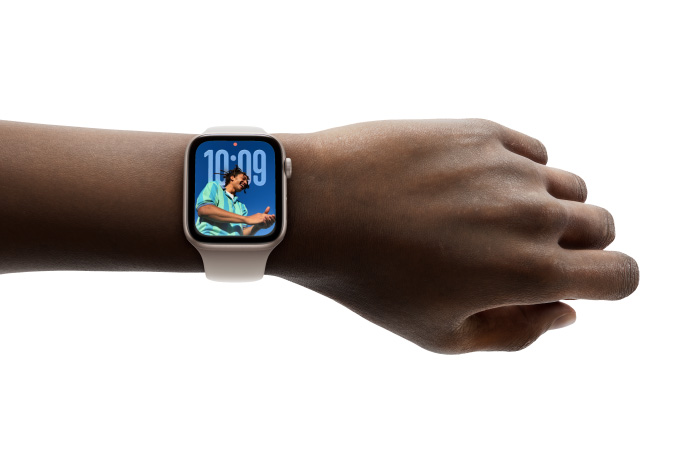 Primo piano di un Apple Watch color oro rosa con cinturino in gomma al polso di una persona, che mostra l’ora nel nuovo design Liquid Glass