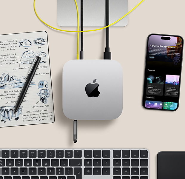 Mac mini, top, silver aluminum, rounded corners, glossy black Apple logo centered, connected by cord, USB stick inserted in USB-C port in front, Studio Display stand partially visible, iPhone, Magic Trackpad, Magic Keyboard with Touch ID, stylus and e-ink sketch pad