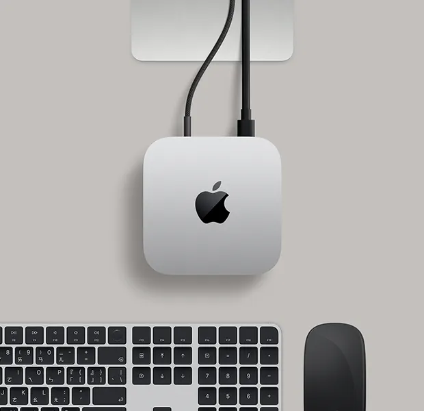 Mac mini 的俯視圖，展示銀色鋁金屬機身、圓角設計，以及機身中央的黑色亮面 Apple 標誌，Studio Display 支架部分可見，旁邊放著含 Touch ID 的巧控鍵盤和巧控板
