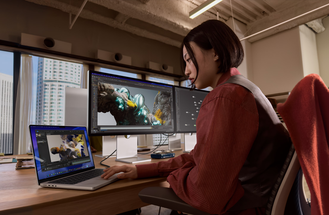 Bir kişi masada oturmuş harici ekrana bağlı MacBook Pro’yla çalışıyor, her iki ekranda da 3D render görünüyor, arkasındaki pencereden binalar görünüyor