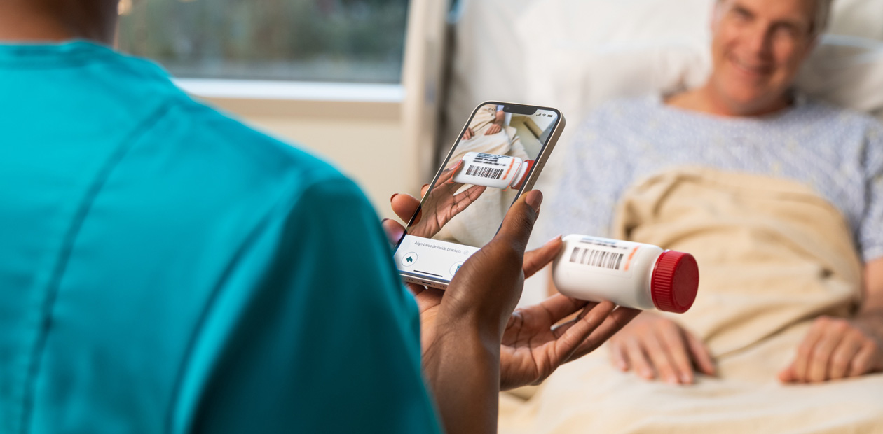 Un’infermiera in camice verde acqua scansiona i medicinali di una paziente con il suo iPhone.