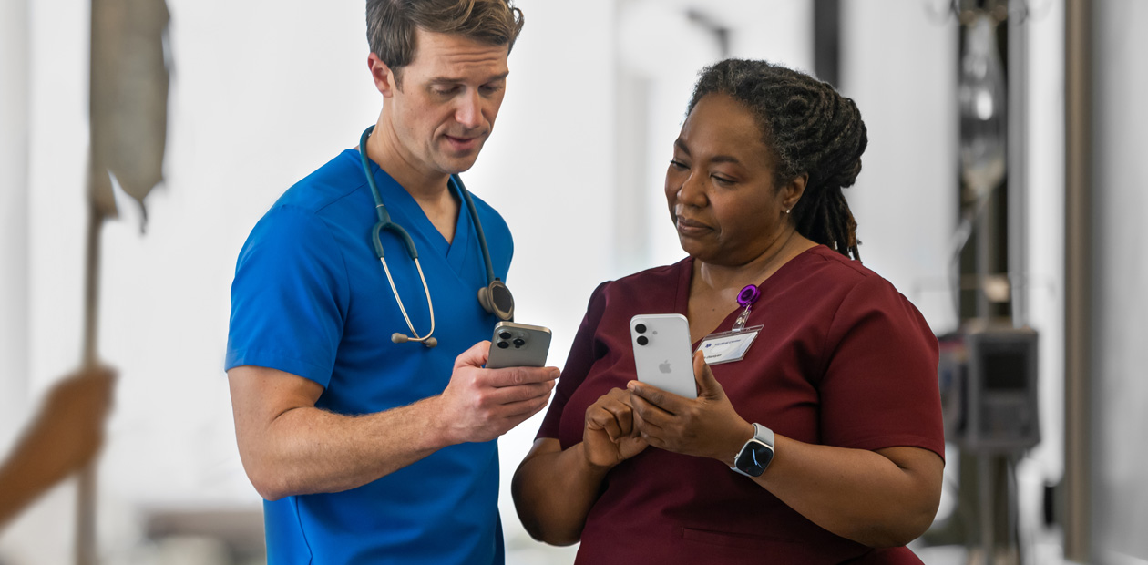 Un medico in camice blu rivolto verso il suo iPhone mostra qualcosa a una medica in camice rosso, anche lei con un iPhone.