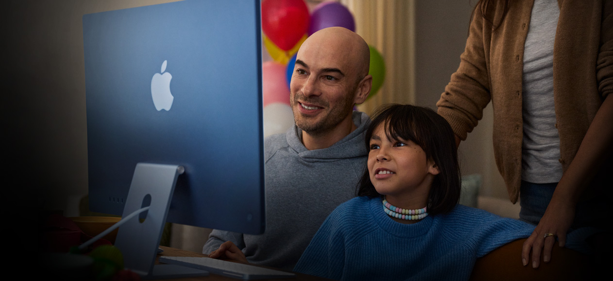 Två personer sitter tillsammans och använder en iMac medan en tredje person står bakom dem och ser på.