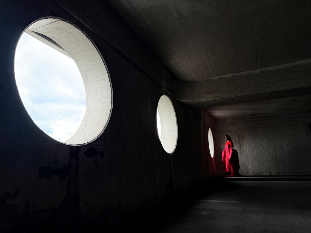 Una donna con un lungo vestito rosso fotografata in piedi in una stanza vuota. Nell’app Foto la lunghezza focale viene regolata a 24 mm, per mettere in evidenza le capacità del sistema di fotocamere Pro